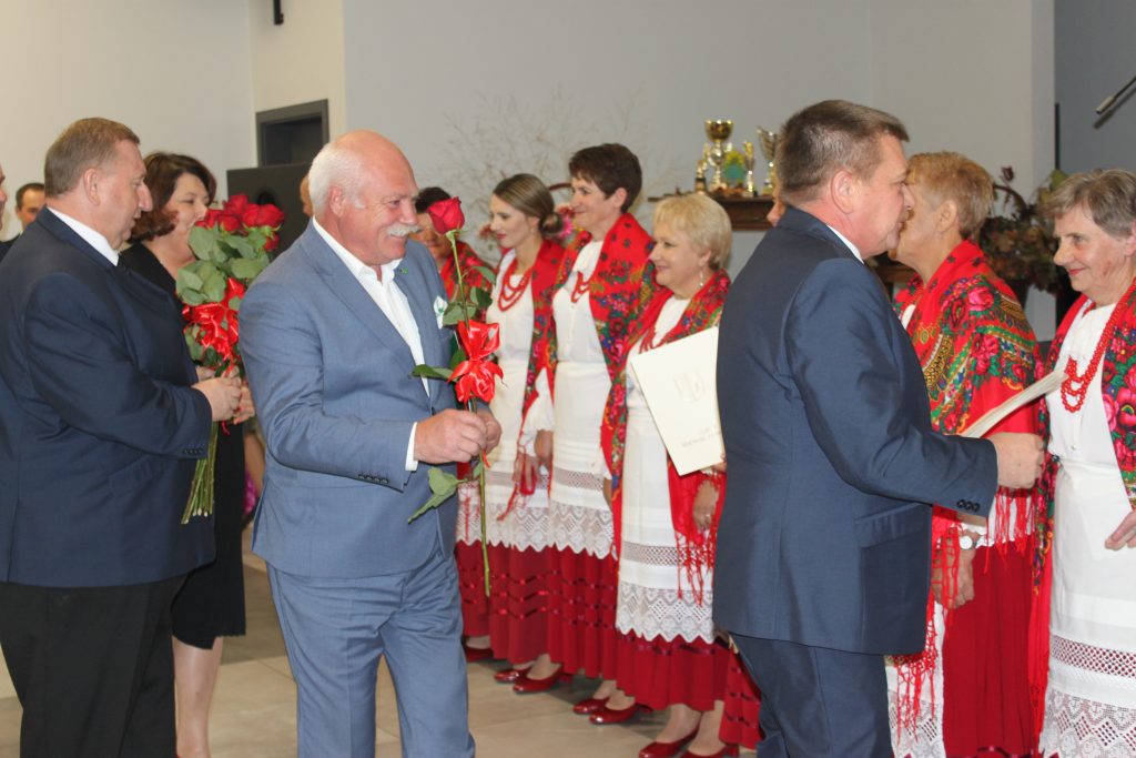 grupa osób stojąca w strojach regionalnych oraz mężczyźni w garniturach wręczający róże