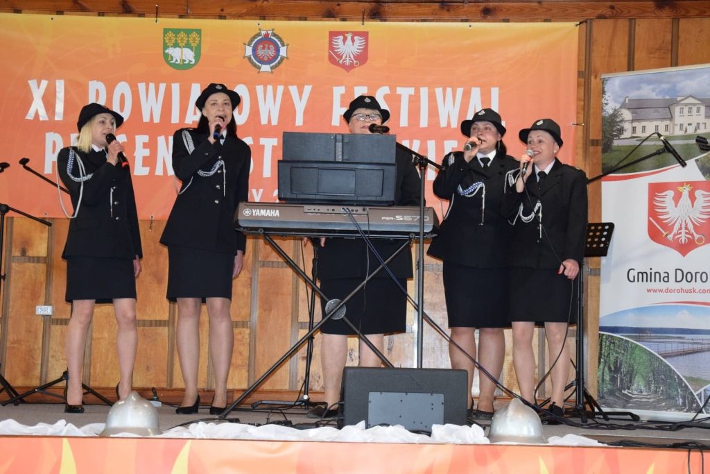 pięć pań w mundurach strażackich śpiewających na scenie