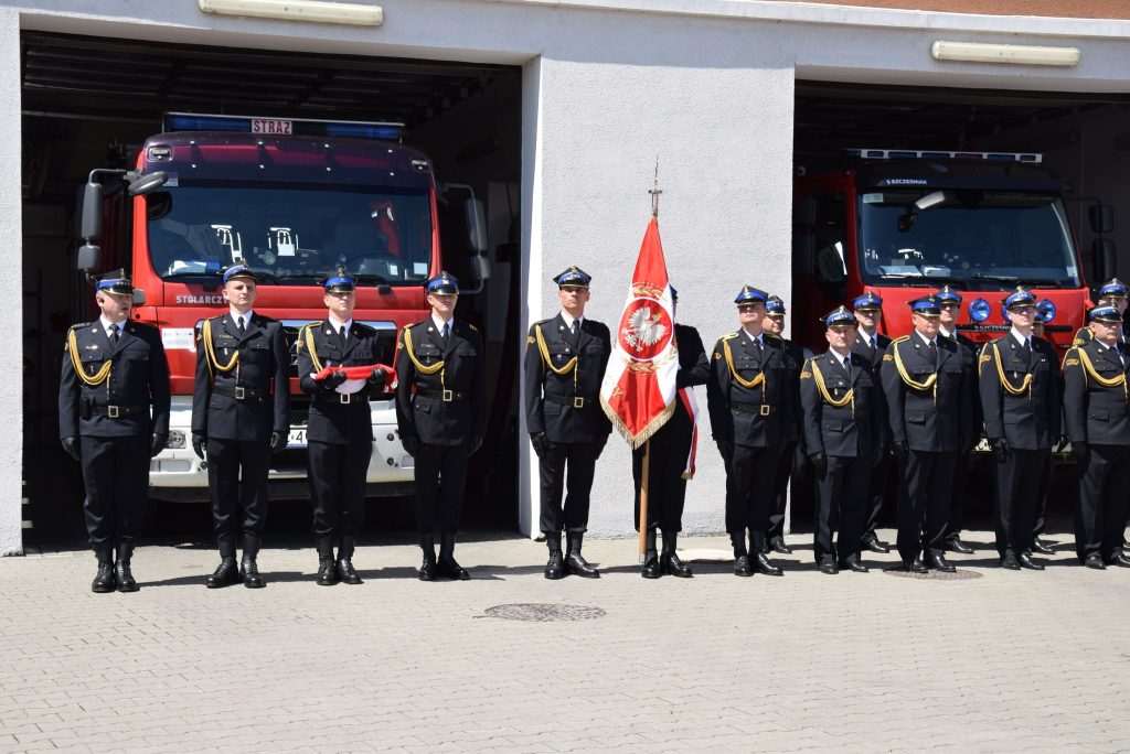 kilkunastu strażaków stojących przed wozem strażackim w mundurach i ze sztandarem