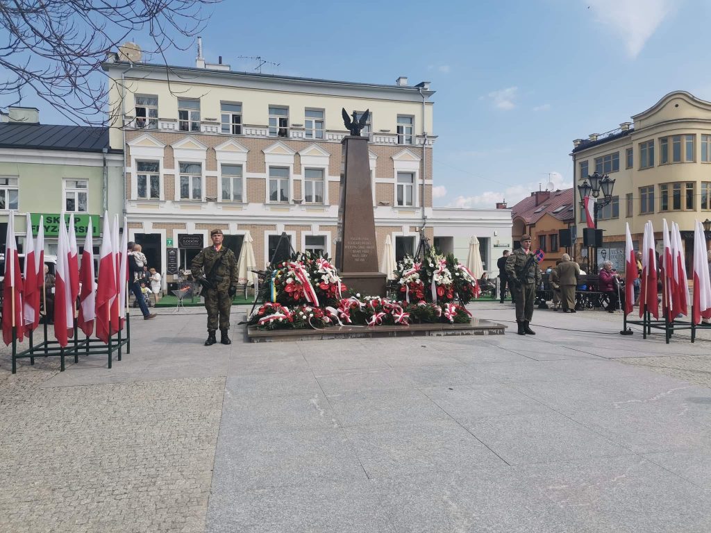 żołnierze stojący przy pomniku obok leżą kwiaty i powiewają flagi Polski