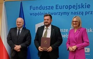Trzy osoby stojące na tle napisu Fundusze europejskie. Mężczyzna trzyma brązową teczkę.