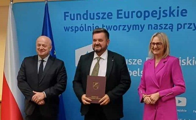 Trzy osoby stojące na tle napisu Fundusze europejskie. Mężczyzna trzyma brązową teczkę. 