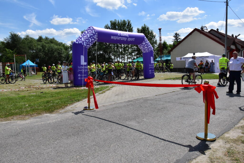 rowerzyści, dmuchana brama w kolorze fioletowym z napisem "z dumą spieramy sport", czerwona wstęga