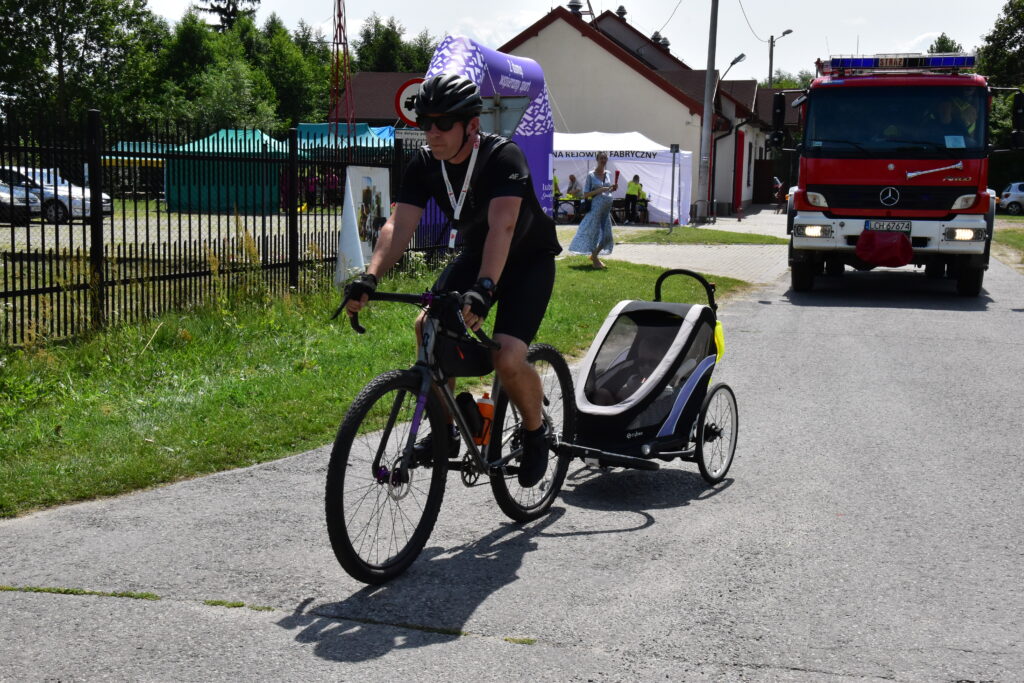 rowerzyści, dmuchana brama w kolorze fioletowym z napisem "z dumą spieramy sport", w tle wóz strażacki