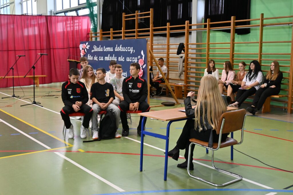 Kilkanaście osób siedzą na krzesełkach w sali gimnastycznej.